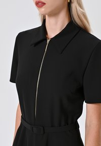 Abito nero con colletto corto, maniche corte e una cerniera frontale con dettagli in oro. La vita è accentuata da una cintura abbinata.