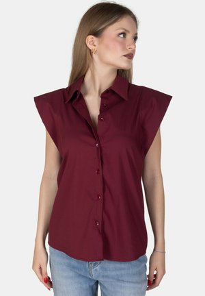 Donna con i capelli lunghi che indossa una camicetta bordeaux senza maniche con bottoni e jeans azzurri chiari, guarda a destra.