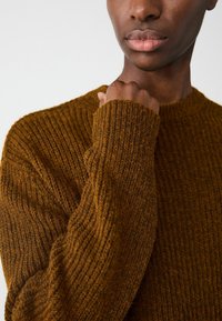 Braun strukturierter Strickpullover mit rundem Halsausschnitt und langen Ärmeln, der ein geripptes Muster und weichen Stoff präsentiert.