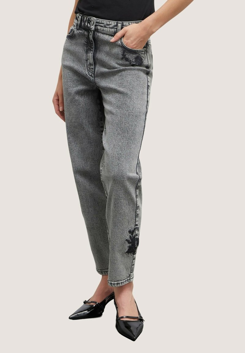 Personne portant un jean gris délavé taille haute avec des imprimés floraux noirs et des talons noirs pointus, debout avec une main dans la poche.