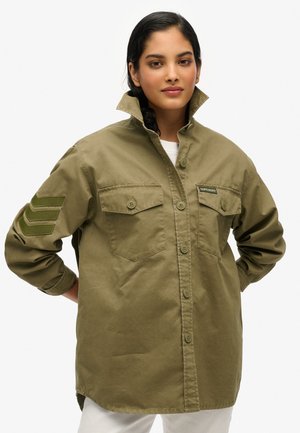Veste chemise vert olive avec deux poches poitrine, fermeture boutonnée et trois rayures texturées sur la manche gauche. Fabriquée dans un tissu léger.