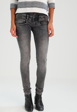Vrouw draagt versleten zwarte skinny jeans, zwarte enkellaarsjes en een wit top met lange mouwen en zwarte horizontale strepen.