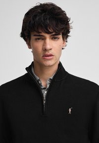 Ung man med mörkt lockigt hår som bär en svart zip-up-tröja över en rutigt skjorta, står mot en enkel ljus bakgrund.