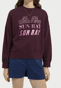 Sudadera color burdeos con gráfico "Sun Ray" en rosa, ajuste holgado, cuello redondo y puños acanalados. Combinada con pantalones cortos azul marino.