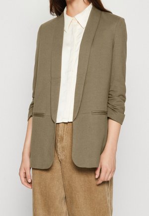Femme portant un blazer olive aux manches retroussées, un chemisier blanc à col, et un pantalon beige en velours côtelé, debout devant un fond clair uni.