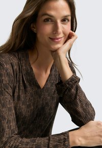 Brown top with vertical black stripes, v-neckline, long sleeves, and a soft texture. Model poses with hand under chin against a neutral background.