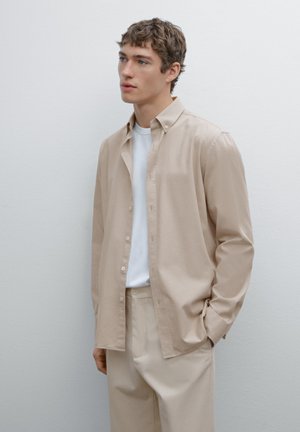 Camicia beige a maniche lunghe con bottoni dal tessuto morbido, abbinata a una t-shirt bianca a collo rotondo e pantaloni beige coordinati, su uno sfondo chiaro.