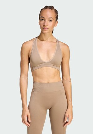 Young woman with braided hair wearing beige sports bra and high-waisted leggings, standing against a plain light background.