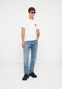 White cotton t-shirt with a red and black graphic design, paired with light blue denim jeans and black chunky sneakers. Black sunglasses worn.