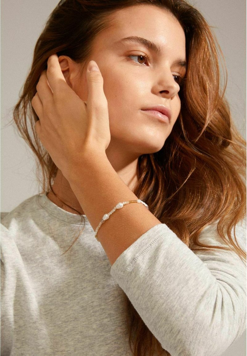 Goldfarbener Armband mit weißen Perlen und kleinen Perlen, getragen an einem Handgelenk, mit einem hellgrauen Langarmshirt, das im Bild sichtbar ist.