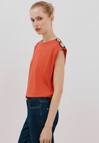 Orange kortærmet top med rund halsudskæring, der har dekorative sort-hvide broderede detaljer på skulderen. Båret med mørke jeans.