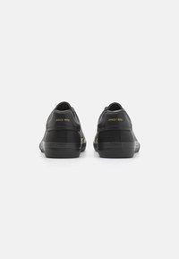 Sapatilhas pretas com tecido texturizado, detalhes suaves e um detalhe em dourado "SINCE 1973" no calcanhar. Design slim com uma silhueta de corte baixo.