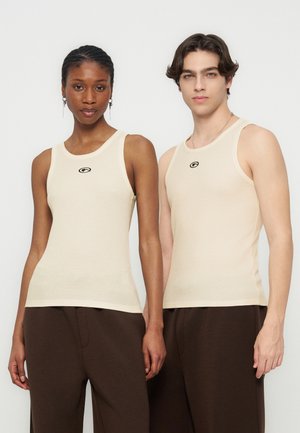 Twee modellen dragen bijpassende beige geribbelde tanktops en donkerbruine broeken, zij aan zij staand tegen een effen achtergrond.