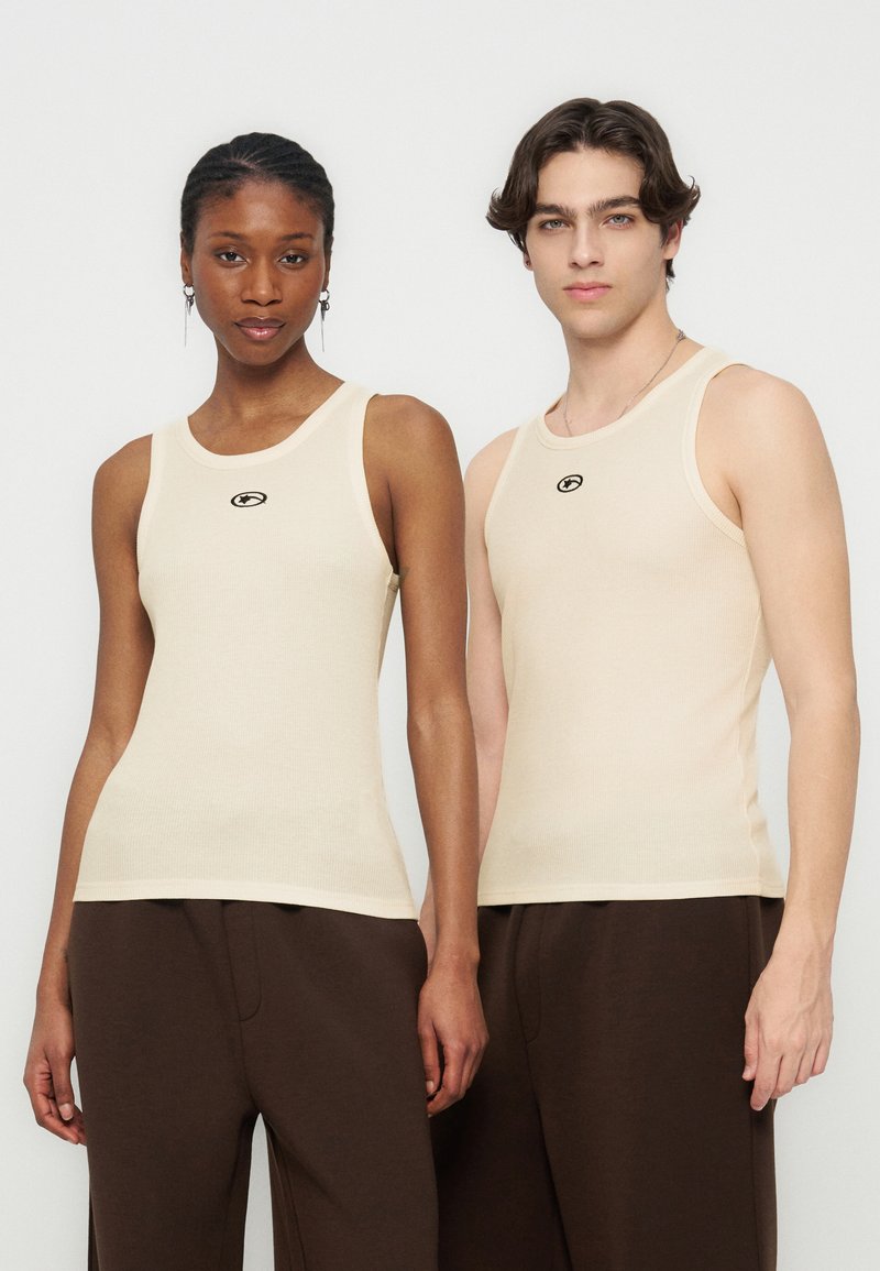 Δύο μοντέλα φορούν ταιριαστά μπεζ ριμππέν τανκ τοπ και σκούρα καφέ παντελόνια, στέκονται δίπλα δίπλα σε απλό φόντο.