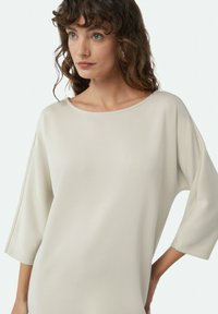 Hellbeige Oversize-Pullover mit weitem Halsausschnitt, Dreiviertelärmeln und glatter Textur. Verfügt über kontrastierende Nähte entlang der Säume.
