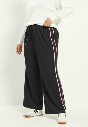 Personne portant un pantalon noir à jambes larges avec des bandes latérales blanches et bordeaux et des baskets blanches avec des accents noirs.