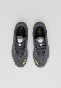 Baskets de sport grises avec une tige en maille, présentant un design texturé, des accents décoratifs jaunes et des lacets noirs contrastants.