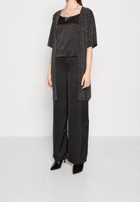 Svart satin topp med en glittrig svart öppen cardigan, matchad med vida svarta byxor och svarta klackskor med spetsig tå. Silverfärgade lagerhalsband syns.