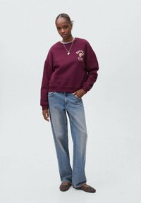 Donna che indossa una felpa color bordeaux con la scritta "Brunch Club", jeans blu larghi, scarpe nere con borchie e collane d'argento, in piedi davanti a uno sfondo semplice.
