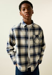 Blau-weiß kariertes Flanellhemd mit Knopfleiste, langen Ärmeln, Brusttasche und legerer Passform über einem weißen T-Shirt.