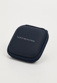 Custodia rigida blu navy con finitura testurizzata, chiusura a zip e logo bianco "TOMMY HILFIGER" sulla parte anteriore. Angoli arrotondati e design compatto.