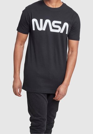 Man wearing a black T-shirt with large white NASA logo across the chest, paired with black pants on a plain gray background.