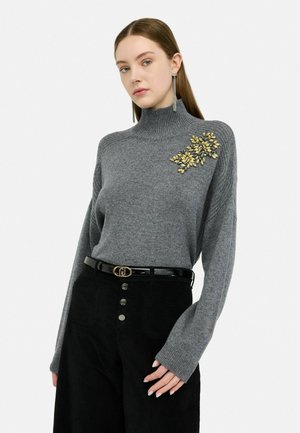Maglione grigio a collo alto con texture a costine e un dettaglio floreale in toni dorati. Abbinato a pantaloni neri a vita alta e una cintura lucida.