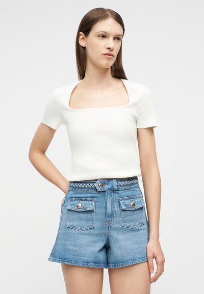 Γυναίκα που φορεί λευκό κοντομάνικο τοπ με τετράγωνο λαιμόκοψη και ψηλόμεσο μπλε τζιν σορτς με μπροστινές τσέπες και πλεκτή ζώνη.