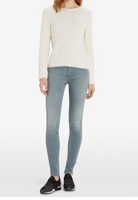 Suéter blanco texturizado, jeans ajustados de color azul claro y zapatillas negras. El suéter tiene un escote redondo y mangas largas. Los jeans se ajustan de cerca.