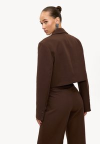 Femme avec un chignon lisse portant un blazer court marron foncé assorti à un pantalon taille haute, regardant par-dessus son épaule, sur un fond blanc uni.