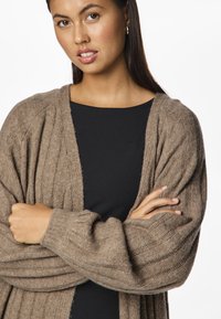 Pruun kootud kardigan, millel on sooniline tekstuur ja avatud esikülg, kantud musta pluusi peal. Omab pikki varrukaid ja laia lõikega disaini.