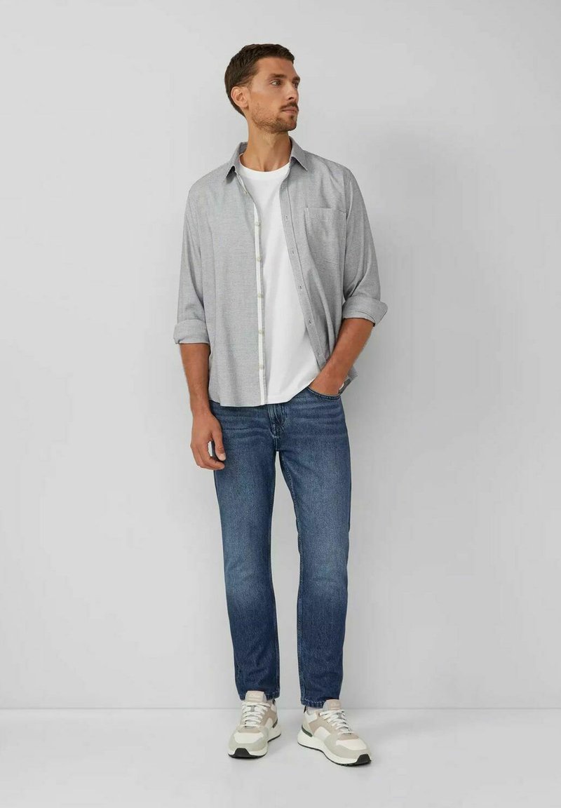 Camisa de botones gris sobre una camiseta blanca, combinada con jeans azules y zapatillas claras. La camisa tiene mangas largas y un bolsillo en el frente.