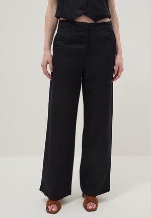 Person trägt schwarze hoch taillierte, weit geschnittene Hose, schwarze kurze Weste und braune offene Absatzschuhe, steht vor einem einfarbigen hellen Hintergrund.