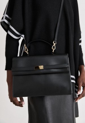 Sac à main en cuir noir avec fermoir doré et bandoulière amovible portée à l'épaule, assortie à un haut à manches rayées noir et blanc et une jupe noire.