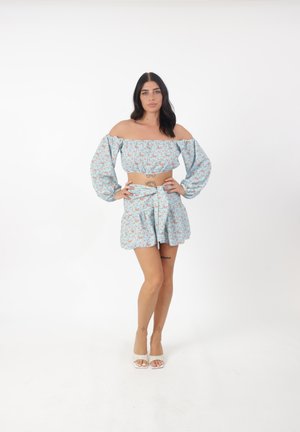 Mujer con cabello largo y oscuro que lleva un crop top azul con flores y hombros descubiertos y una falda cruzada a juego, de pie con las manos en las caderas sobre un fondo blanco.