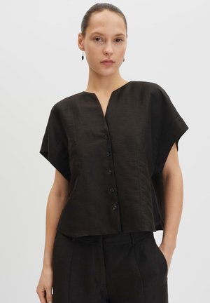 Femme aux cheveux tirés en arrière, portant un chemisier noir à manches courtes boutonné et un pantalon noir, debout avec une main dans la poche.