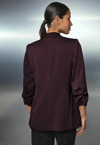 Donna di spalle che indossa un blazer viola scuro con le maniche arrotolate e orecchini a cerchio dorati, capelli raccolti in una coda di cavallo.