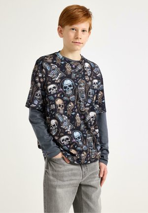 Rødharret dreng iført lagdelt grå langærmet skjorte og sort T-shirt med flerfarvet kranium og skateboard grafik, stående med hænderne i lommerne.