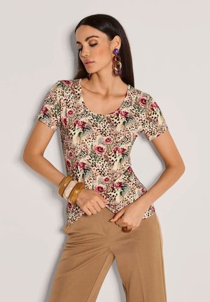 Vrouw in een kort mouwende bloeiende en luipaardprint top, beige broek, paarse oorbellen en chunky bruine armbanden met handen op de heupen.
