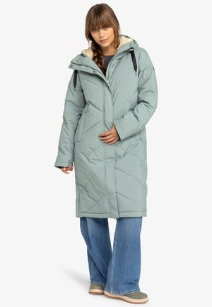 Padded long coat in light green. Features a zip closure, fur-lined hood, and diamond quilted design. Paired with wide-leg jeans and boots.