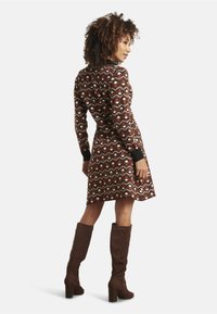 Braun gemustertes Kleid mit geometrischen Formen, mit langen Ärmeln und ausgestellt geschnittenem Saum. Kombiniert mit kniehohen braunen Stiefeln.