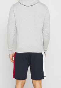 Felpa grigia con cappuccio e coulisse, abbinata a pantaloni corti blu scuro con strisce laterali rosse e bianche. Composizione in cotone, vestibilità casual.
