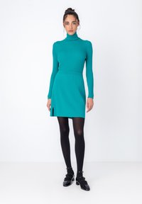Zelena rebrasta turtleneck majica v kombinaciji z usklajeno zeleno mini krilo z sprednjim razporkom, oblečena z črnimi pajkicami in čevlji s peto.