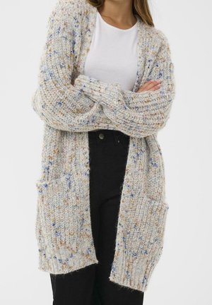 Femme portant un long cardigan en maille épaisse avec des taches multicolores sur un haut blanc et un pantalon noir, bras croisés.