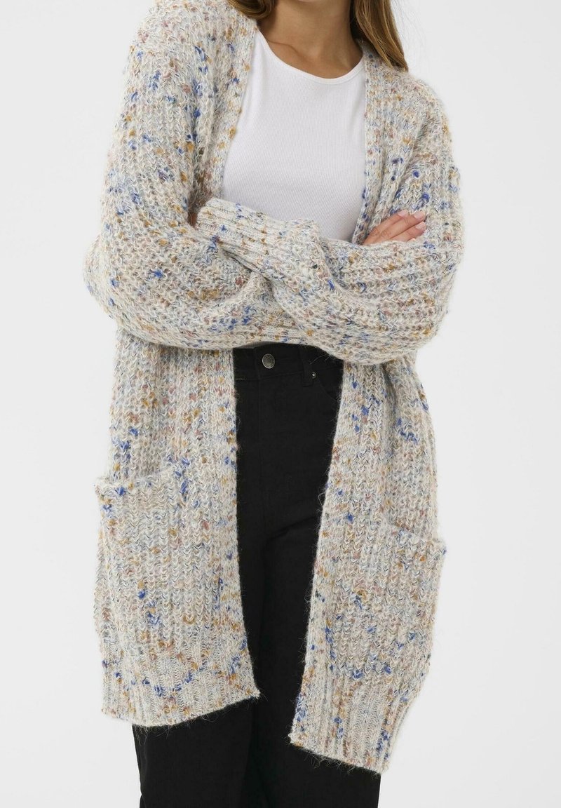 Femme portant un long cardigan en maille épaisse avec des taches multicolores sur un haut blanc et un pantalon noir, bras croisés.