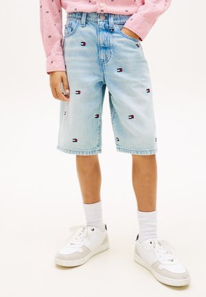 Dziecko w jasnoniebieskich szortach jeansowych z drobnymi logo flag, białych trampkach, białych skarpetkach oraz różowej bluzce z długim rękawem ozdobionej wzorami.