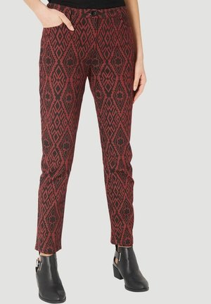 Mujer vestida con pantalones de patrón geométrico rojo y negro, botas negras hasta el tobillo y una camiseta negra sobre un fondo neutro.