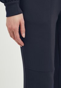 Marinblå idrottsbyxor med en jämn textur, med fickdetalj p�å låret och elastiska muddar vid anklarna. Närbild av en hand på tyget.