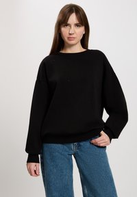 Czarna bluza z okrągłym dekoltem, luźny krój i długie rękawy. Posiada subtelne zdobienia na przodzie i jest zestawiona z jasnoniebieskimi dżinsami.