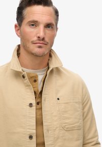 Beige, gekraagde jas met knoopsluiting en een enkele borstzak. Draag het over een bruine henley van textiel. Schoon ontwerp, veelzijdig uiterlijk.